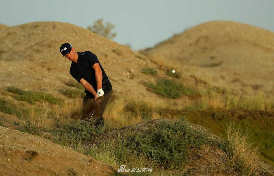 球童为球员擦拭高尔夫球，背景是典型的沙漠植被与沙坑，两人正为下一杆关键击球做细致准备。